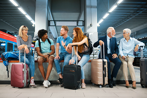 Closeup front view of group of young adults and a senior couple waiting for a train ride at an underground railroad station. They are sitting on a bench and talking.