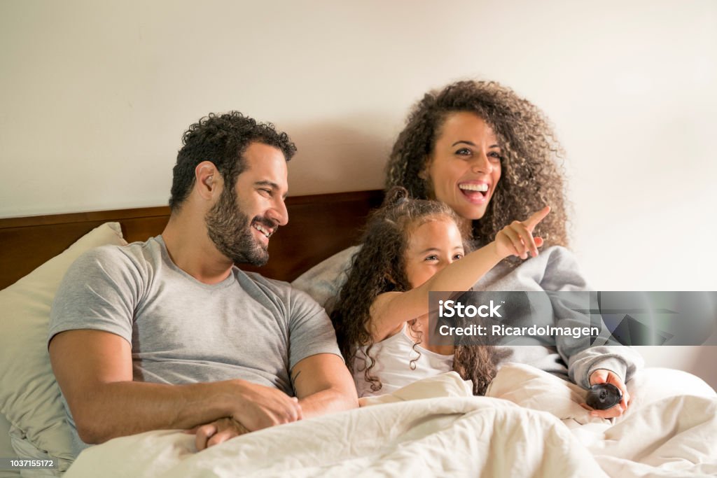 Dad, mom and daughter in bed Interracial family, lying in bed, smiling as they watch television, in the privacy of their home Bed - Furniture Stock Photo