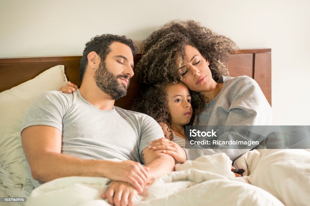 Dad, mom and daughter in bed Interracial family, lying on the bed, trying to sleep, while the little girl watches tv 2-3 Years Stock Photo