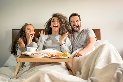 Interracial family, lying on the bed, with a surprised face, while they have breakfast on a tray on the bed and point something out