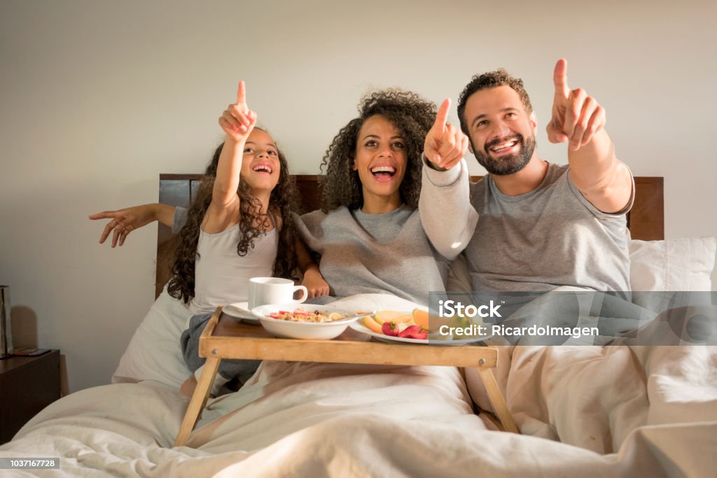 Family having breakfast in bed Interracial family, lying on the bed, with a surprised face, while they have breakfast on a tray on the bed and point something out 6-7 Years Stock Photo