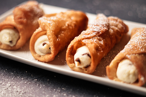 Feshly made cannoli. Crispy shells are filled woth luscious ricotta and marscapone cheese with a hint of lemon zest and mini chocolate chips. Served on a white platter with dusted powedered sugar.