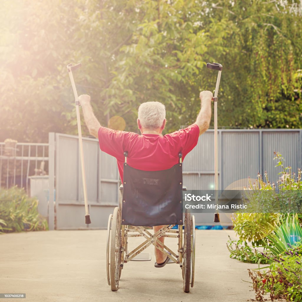 Senior in a wheelchair cheering victoriously Back View of an older, handicapped man, arms and crutches raised in happiness, sunflare Celebration Stock Photo