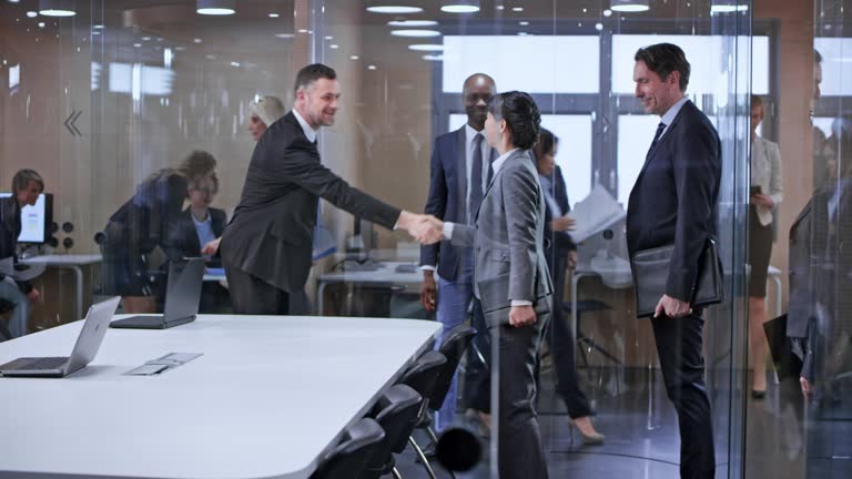 Wide dolly shot of a group of business people entering the glass conference room and shaking hands with the other team. Shot in Slovenia.