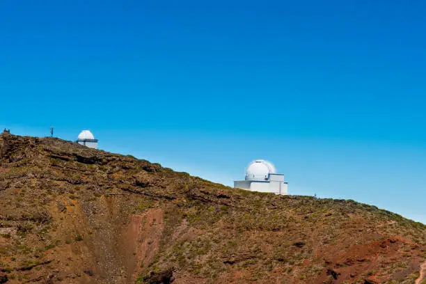 Telescopes in bright daylight behind reddish rocks below a clear blue sky