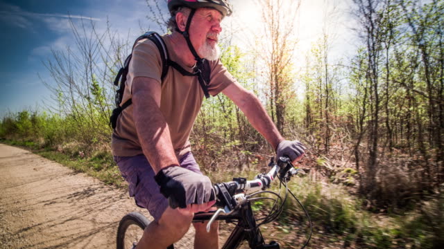 ドイツで70歳のサイクリングをしたシニアマン。レンズに入ってくる太陽フレアと暖かい日光で照らされたバック。彼はサイクリングヘルメットをかぶっている。