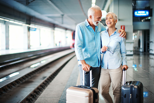 Closeup of an early 60's couple waiting for a train at a train station. They are standing on the platform with suitcases on the side, the man emraced his wife, they are talking and laughing. Copy space on the left.