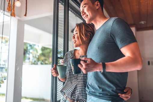 Smiling couple looking through window while holding mugs at home. Young man and woman are spending leisure time together. They are standing in living room.