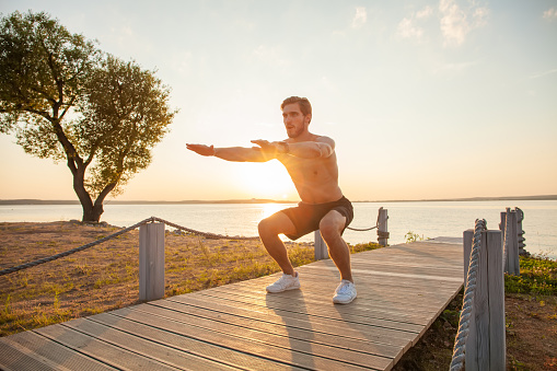 Fitness man training air squat exercise on beach outside. Fit male exercising gym outside. Young handsome caucasian male fitness model and instructor outdoors