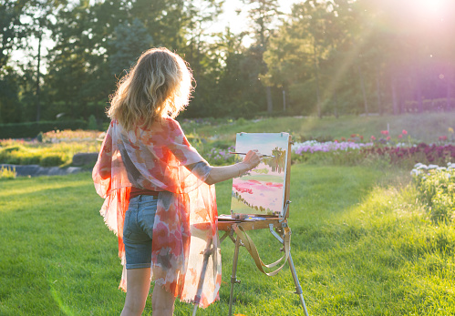 A young woman artist holds a brush and paints a picture on an easel in the rays of the sunset. The painter paints oil paintings in the garden at sunset