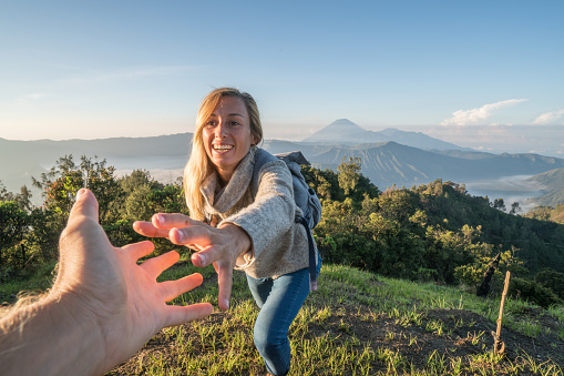 Young woman hiking, pulls out hand to reach the one of teammate. A helping hand to reach the mountain top. Bromo volcano region in Indonesia, Asia