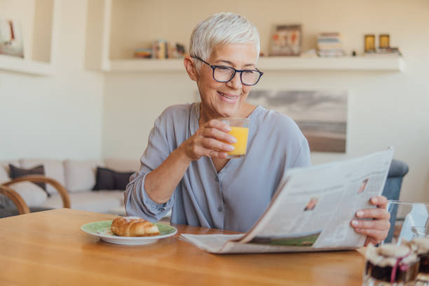 senior mujer bebiendo jugo de naranja en casa - mujer leyendo periodico fotografías e imágenes de stock
