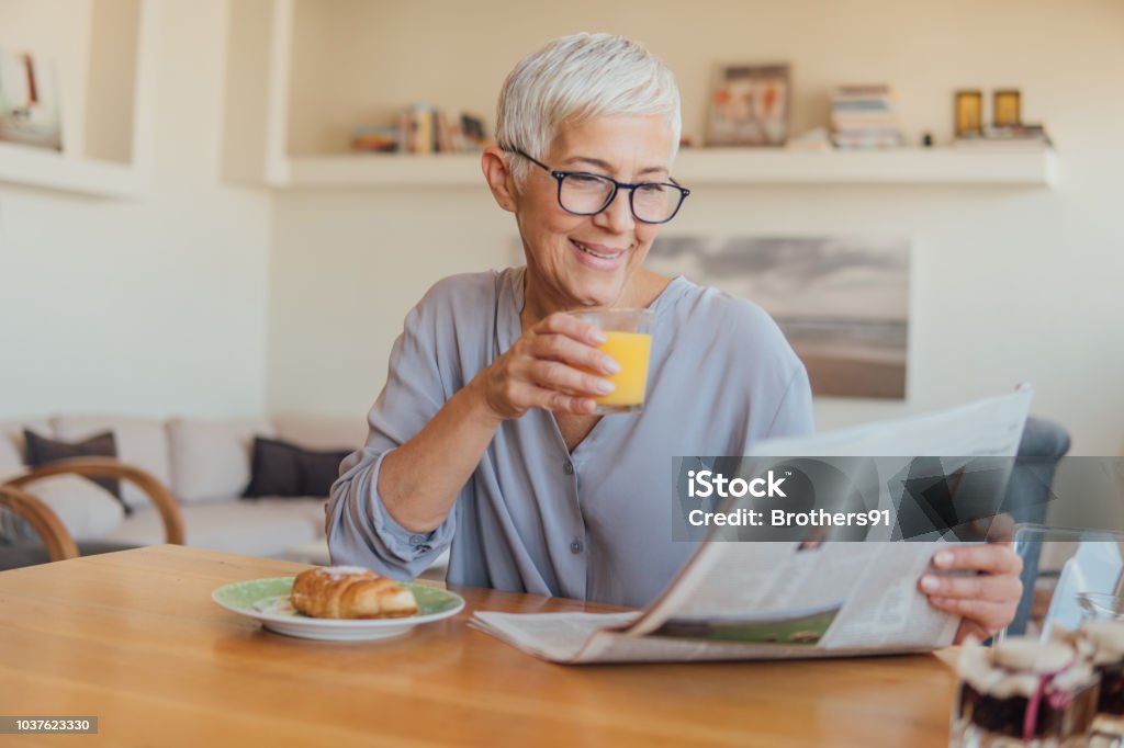 Senior woman drinking orange juice at home Elderly woman holding newspapers while having breakfast at home Newspaper Stock Photo