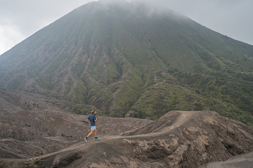 Young man running on cross country dirt trail in Indonesia on volcanoes with dramatic landscape. People travel adventure fun concept