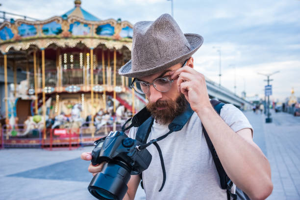 istock bearded young man in hat and eyeglasses using digital camera in amusement park 1037670444