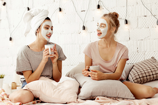 Portrait of beautiful smiling girls with cosmetic cleansing masks sitting in bed drinking hot coffee and having open dialogue. Cozy atmosphere and lovely interior in light colours