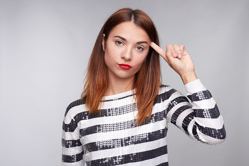 Unhappy exhausted mixed raceds female imitates gun shoot, makes suicide gesture, touches temple, has pure skin, curves red lips, has straight ginger hair, isolated over gray studio wall background.