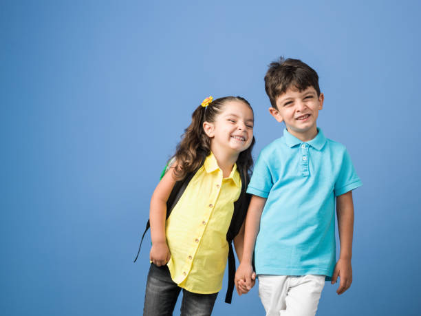 Portrait Of Schoolboy And Schoolgirl Holding Hands In Front Of Blue Background Portrait of little schoolgirl and schoolboy carrying school bags in front of blue background. Boy is wearing a turquoise t-shirt and beige pants while girl is wearing a yellow blouse and black jeans. Shot in studio with a medium format camera. blue brothers stock pictures, royalty-free photos & images