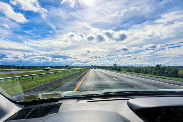 Driver car point of view perspective view looking through the windshield of a westbound vehicle speeding in the left lane on the New York State Thruway multi-lane interstate highway expressway on a bright, sunny September summer day under a puffy, late afternoon cloudscape. Several semi-truck tractor trailer trucks are visible up ahead hauling freight in both directions. New York State Thruway Interstate Route 90 surrounded by the Montezuma National Wildlife Refuge - located about halfway between Syracuse, NY and Rochester, NY.