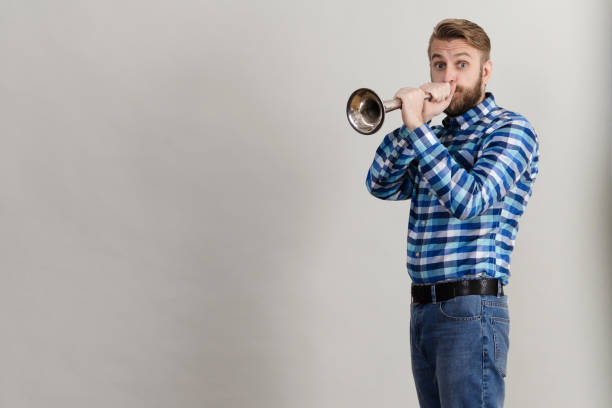 Funny bearded man in a plaid shirt blowing into the copper horn Funny bearded man in a plaid shirt blowing into the copper horn isolated on grey background man trumpet stock pictures, royalty-free photos & images