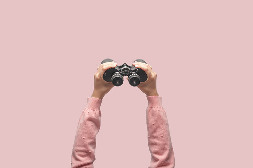 Woman with binoculars on retro pink background, looking through binoculars, journey, find and search concept.
