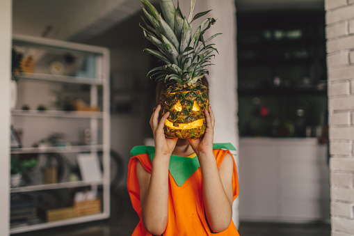 Boy dressed as a pumpkin covering his face with a carved pineapple on a Halloween