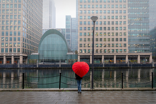 Woman with red, heart shaped umbrella stands in London Canary Wharf on a rainy autmun day