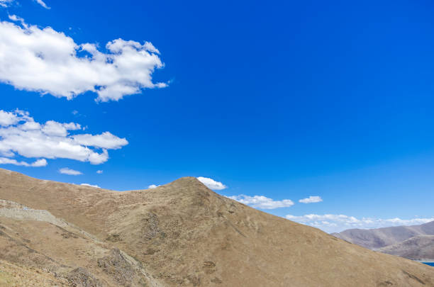 Desert hillside with bushes and tall barren mountains beyond under bright blue sky.