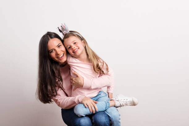 A small girl with crown headband and her mother sitting in a studio. A small girl with crown headband sitting on her mother's lap. A studio shot on a white background. dark pink dress stock pictures, royalty-free photos & images