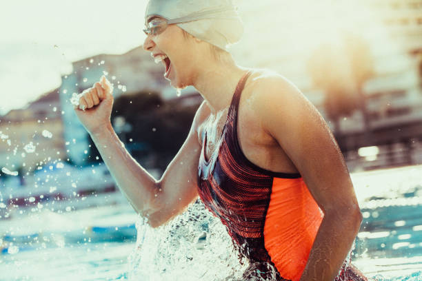 Excited female swimmer celebrating victory Excited female swimmer with clenched fist celebrating victory in the swimming pool. Woman swimmer cheering success in pool wearing swim goggles and cap. sportsperson stock pictures, royalty-free photos & images
