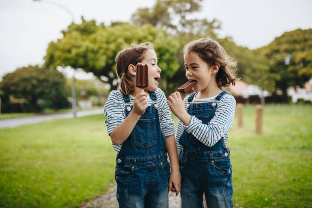 Twin sisters enjoying eating candy icecream Two little twin sisters in identical clothes standing outdoors and eating chocolate ice cream candy. Two little girls enjoying eating candy icecream outdoors. twin stock pictures, royalty-free photos & images