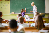 Senior female teacher teaching schoolgirl in front of blackboard at elementary school.