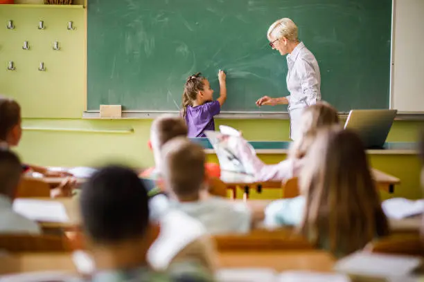 Happy female teacher talking to schoolgirl while writing on blackboard in the classroom.