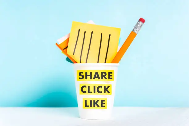 Photo of Share click like words. Paper cup with post-it notes and pencil on blue background over desk.