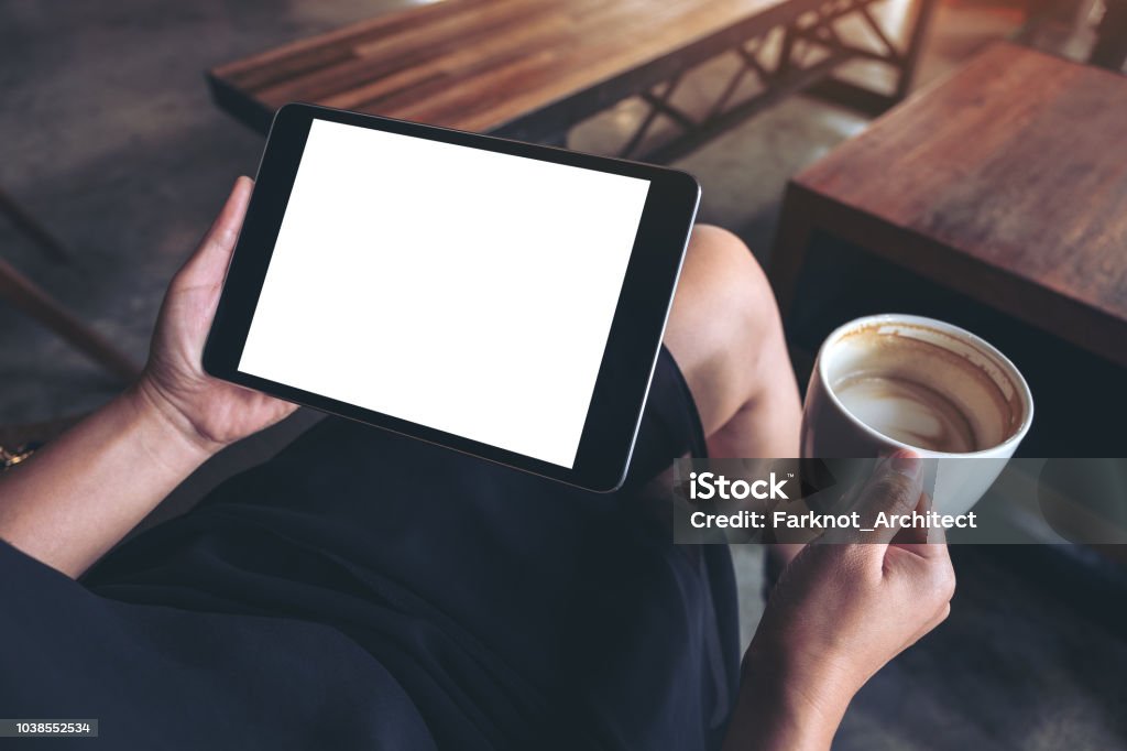 Mockup image of woman's hands holding black tablet pc with blank white desktop screen while sitting and drinking coffee in cafe Above Stock Photo