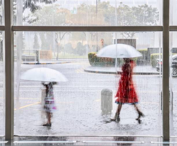 chuva torrencial em tropical que singapura vista através de uma janela listado com água, como uma menina jovem e mulher andam lá fora com a correspondência de guarda-chuvas brancos, seu movimento borrado. - blurred motion clothing mother offspring - fotografias e filmes do acervo