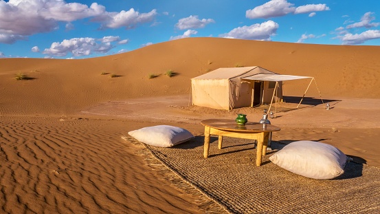 The curves, lines and dramatic landscape of the Sahara Desert, with a bright blue sky and fluffy white clouds. A campsite is set within the dunes, including an outdoor sitting area with carpet, table and cushions, and a tent in the background.