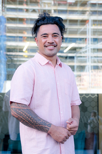 Portrait of proud smiling man looking at camera. Low angle view with reflection of urban construction site in background. Male is of Samoan ethnicity and has tattoos and facial hair moustache.