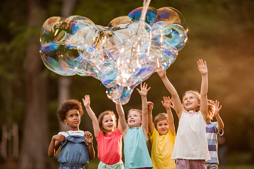 Group of happy kids having fun with heart shaped rainbow bubbles in springtime at the park. The heart made of bubbles is real.