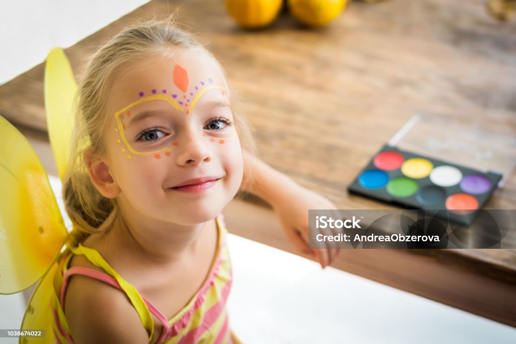 Linda niña con pintura de cara, sentado en una mesa, mirando a la cámara sonriendo. Fondo familia estilo de vida fiesta o Carnaval de Halloween. Pintura de la cara y vestirse. - Foto de stock de Niño libre de derechos