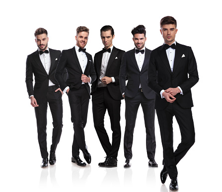 group of five grooms in black tuxedoes with leader fixing sleeves in front, standing on white background, full body picture