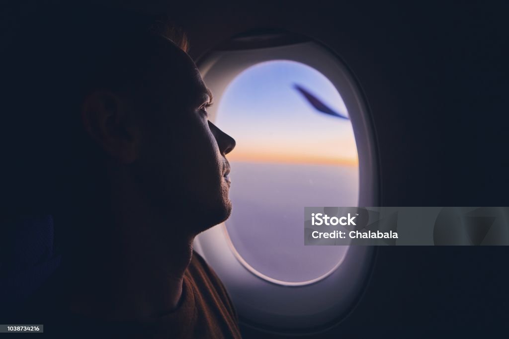 Man traveling by airplane Silhouette of young man traveling by airplane. Pensive passenger looking through window during flight at sunrise. Airplane Stock Photo