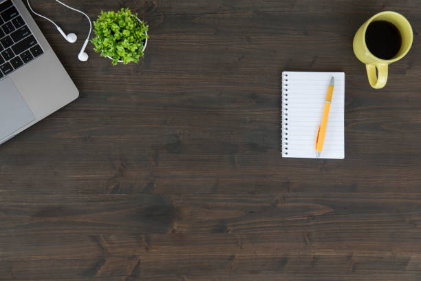 Top view of wooden table with laptop, plant, headphones, coffee cup and note pad with pencil.