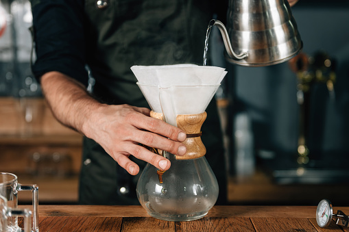 Barista pouring hot water from kettle into white filter for drip coffee. Barista working in coffee shop, wearing dark uniform.