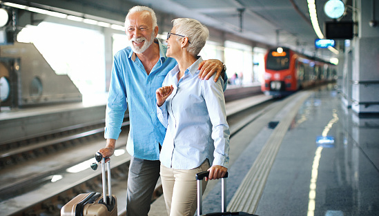 Closeup of an early 60's couple waiting for a train at a train station. They are standing on the platform with suitcases on the side, the man embraced his wife, they are talking and laughing. Copy space on the right.