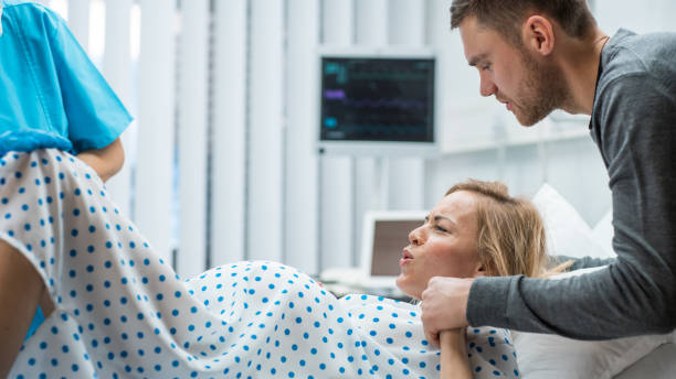 in the hospital, woman in labor gives birth, obstetricians and doctors assist, her husband supports her by holding hand. modern maternity/ delivery ward with professional midwives. - labour room imagens e fotografias de stock