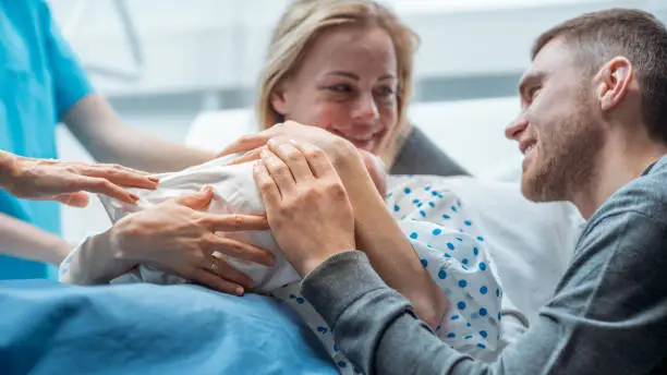 Photo of In the Hospital Midwife Gives Newborn Baby to a Mother to Hold, Supportive Father Lovingly Hugging Baby and Wife. Happy Family in the Modern Delivery Ward.
