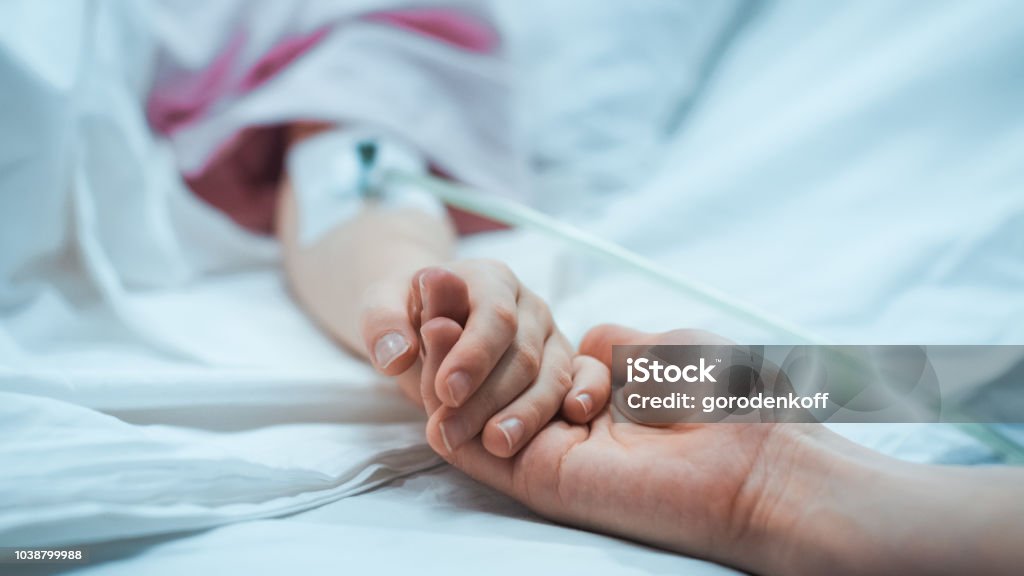 Recovering Little Child Lying in the Hospital Bed Sleeping, Mother Holds Her Hand Comforting. Focus on the Hands. Emotional Family Moment. Hospital Stock Photo