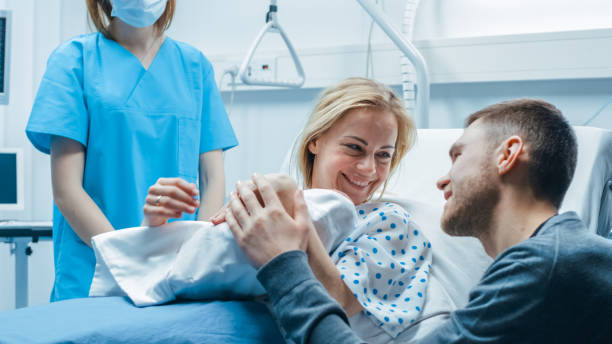 in the hospital midwife gives newborn baby to a mother to hold, supportive father lovingly hugging baby and wife. happy family in the modern delivery ward. - new childbirth new life love imagens e fotografias de stock