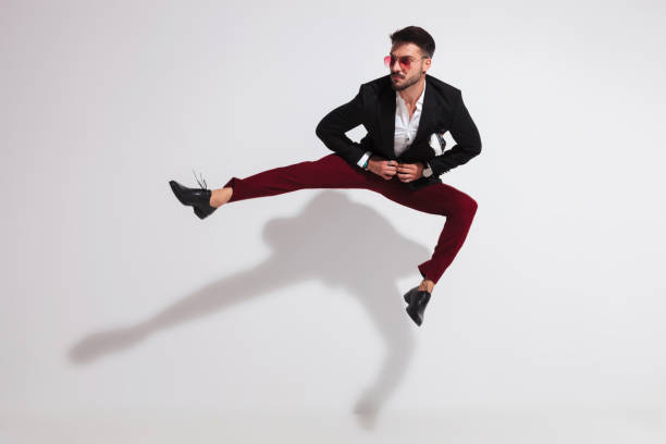 jumping young man parting his legs and unbuttoning black suit jumping young man parting his legs and unbuttoning black suit while looking to side near a white wall legs against the wall stock pictures, royalty-free photos & images